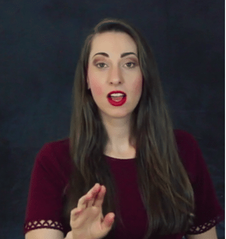 Eine Frau mit langen braunen Haaren, rotem Lippenstift und einem angenehmen Gesichtsausdruck hebt ihre linke Hand in einer Stopp-Geste.