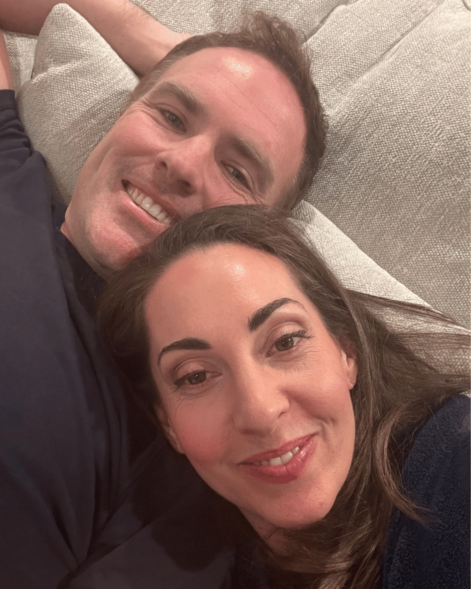 A couple lie head to head on a couch, both smiling at the camera.