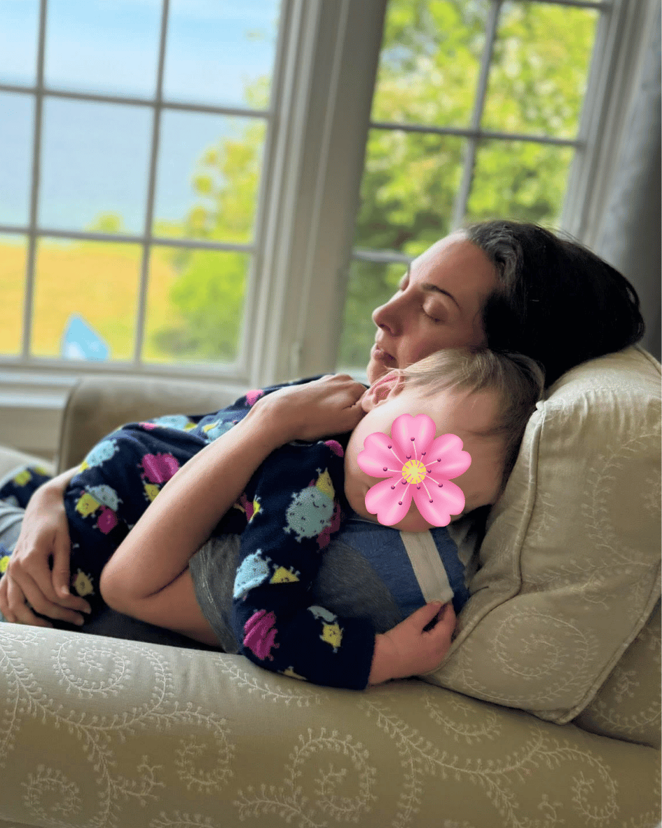 A woman naps on a sofa holding a young child, sunlit windows behind them.