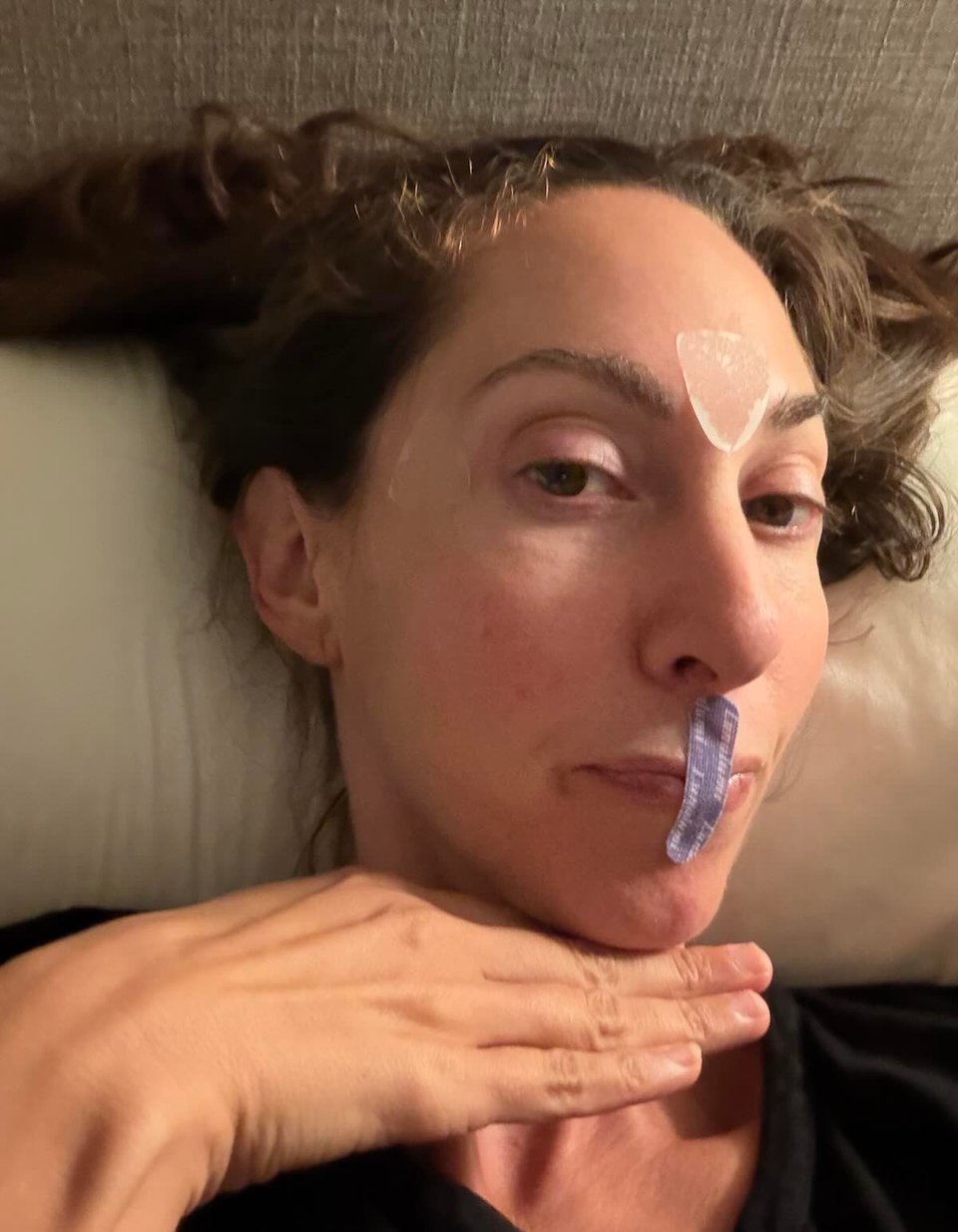 A woman lies on a pillow at night wearing a strip of mouth tape and under-eye patches.