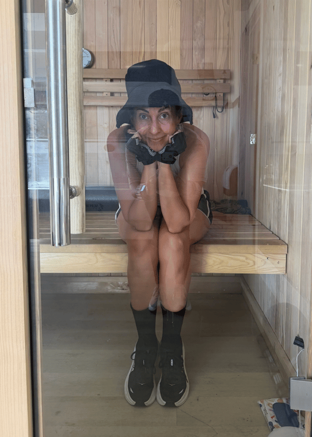 A woman sits in a wooden sauna wearing a wide-brimmed sauna hat, knees pulled up, smiling.
