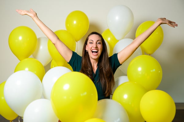 Vanessa Van Edwards with arms raised excitedly among yellow balloons