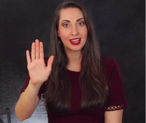 Eine Frau mit langen braunen Haaren, rotem Lippenstift und einem angenehmen Gesichtsausdruck hebt ihre linke Hand in einer Stopp-Geste.