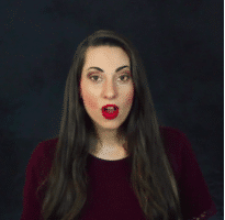 Eine Frau mit langen dunklen Haaren und auffälligem rotem Lippenstift schaut überrascht, ihr Mund ist zu einem „O“ geformt, vor einem dunklen Hintergrund