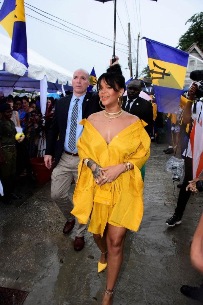 Rihanna walking through a crowd as she holds her micro purse in front of her. with her arms corssed