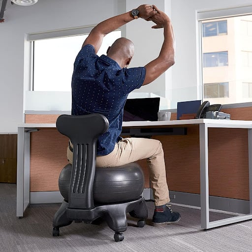 A man is at work sitting on a yoga balance ball doing a side stretch as an office exercise