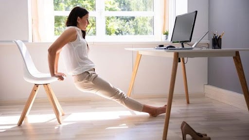A woman is working from home doing tricep dips in her chair as an office exercise