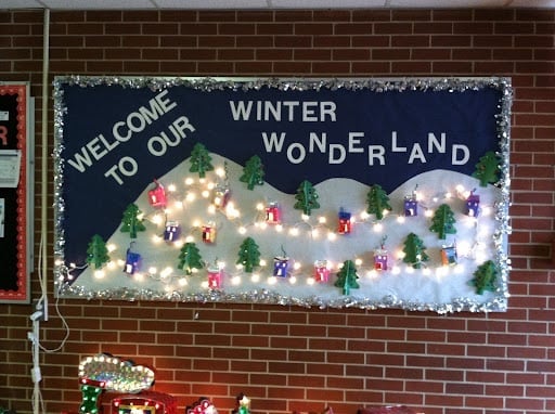 An image of a bulletin board idea for the holidays with the title "welcome to our winter wonderland". It has fairy lights strung on it, as well as cotton for the snow, little trees, and little houses to make it look like a winter scene.