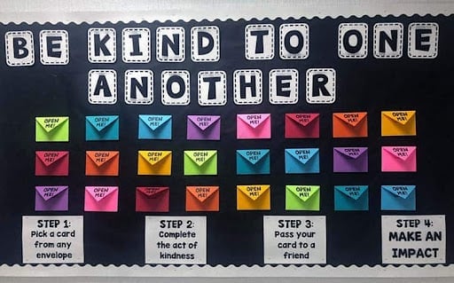 An image of a bulletin board idea with the title "be kind to one another". It has a bunch of colorful envelopes with random acts of kindness in each one, so that employees can take on or two and carry out the random act of kindness, before putting it back to pass it on to someone else.
