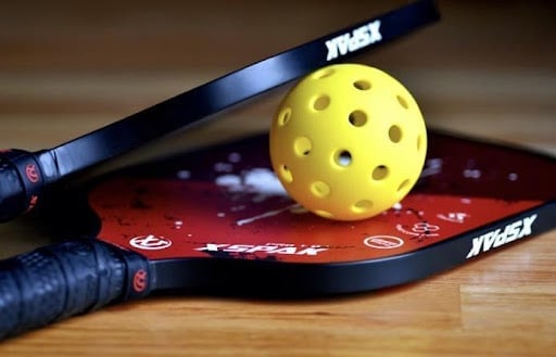 An image of two pickleball paddles and a wiffle ball. It’s a great party game that is a combination of tennis, badminton, and ping pong, played on a smaller court with a lower net and a paddle instead of a racket.
