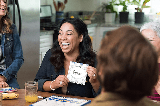 A woman holding up a square card that says beaver on it while laughing. She is playing a game called Telestrations with friends, which is similar to Pictionary, and is a hilarious drawing game where players sketch out clues and try to guess what their teammates have drawn.