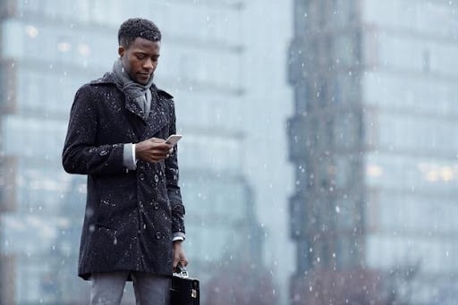 An example of a man changing his office wardrobe for the seasons. He is walking in the snow looking at his phone wearing layers. This is something to consider as part of office dress code. 